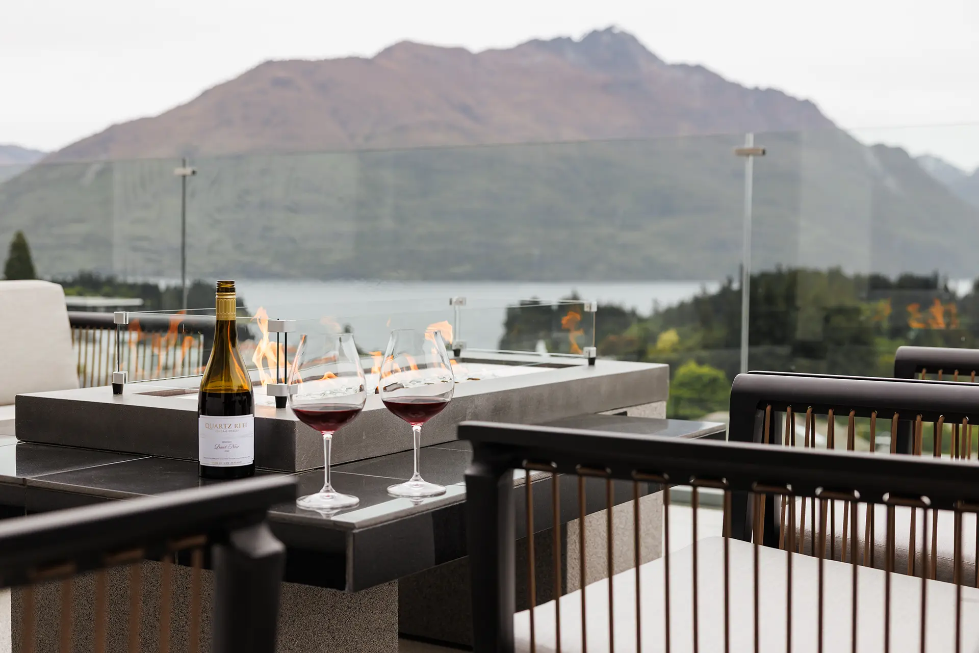 The Carlin Hotel - Two glasses of red wine and a wine bottle on a table with a glass-enclosed fire pit overlooking mountains and a lake.