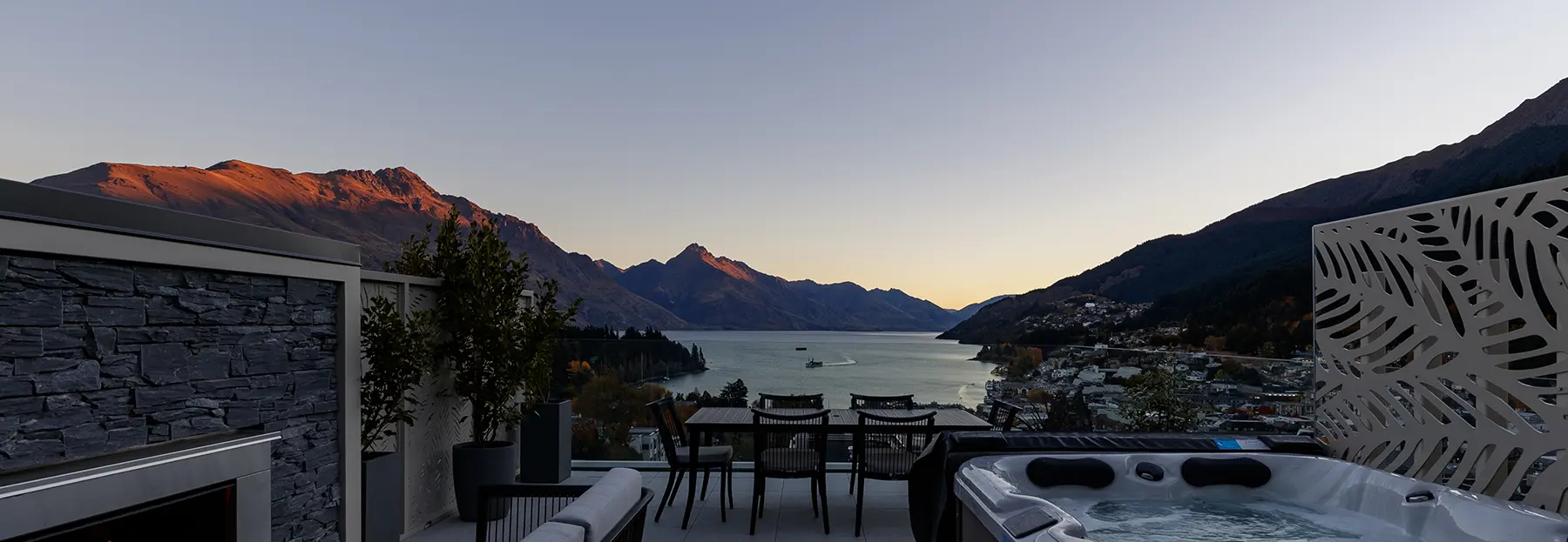 The Carlin - Terrace with jacuzzi, seating area, and dining table overlooking a lake and mountains at sunset.