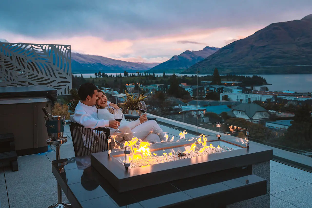 Couple in white clothing sitting close together on a balcony with wine glasses by a modern outdoor fire pit, overlooking a lake and mountains at sunset.