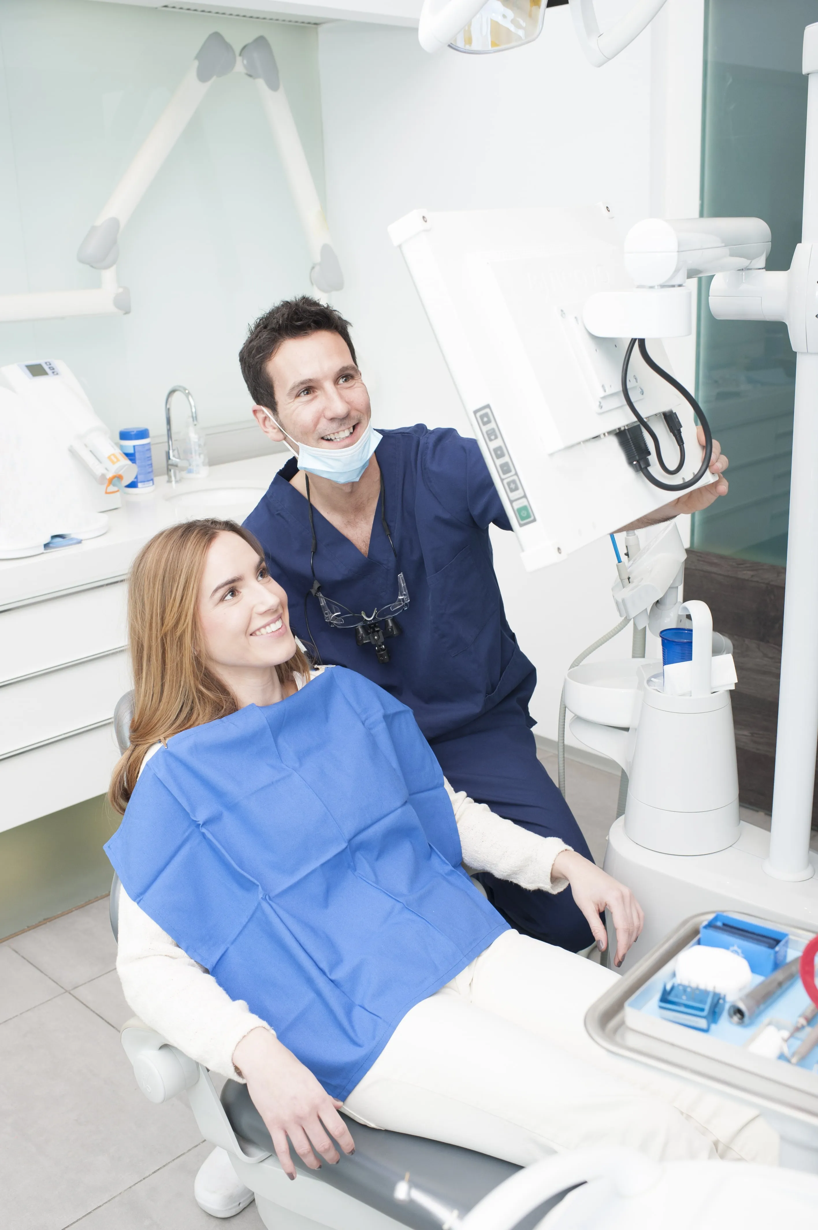 Dentist wearing navy scrubs and a face mask talks to a smiling female patient seated in a dental chair while showing an image on a monitor.