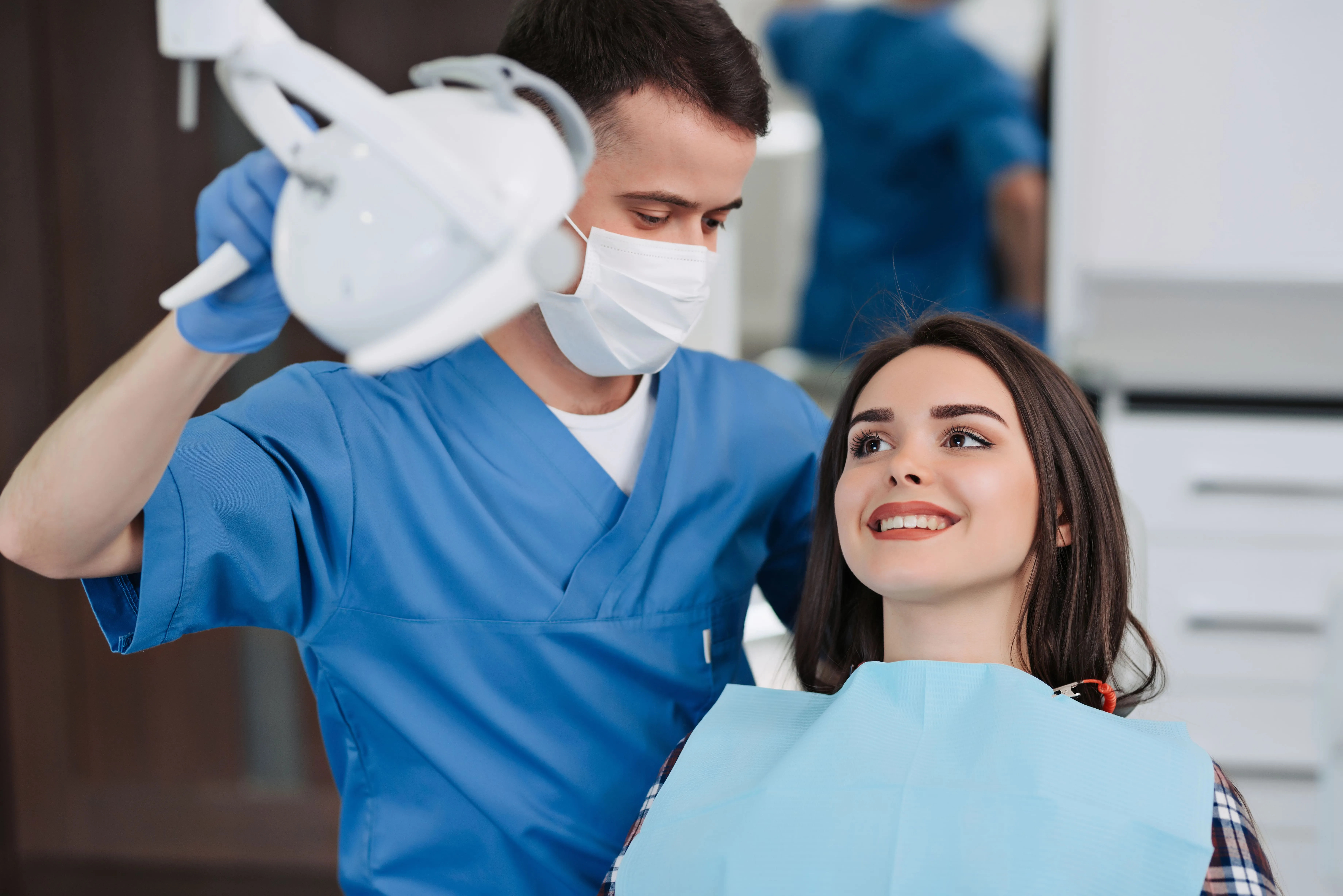 Dentist in blue scrubs and mask adjusts dental light while smiling female patient with a dental bib sits in the chair.