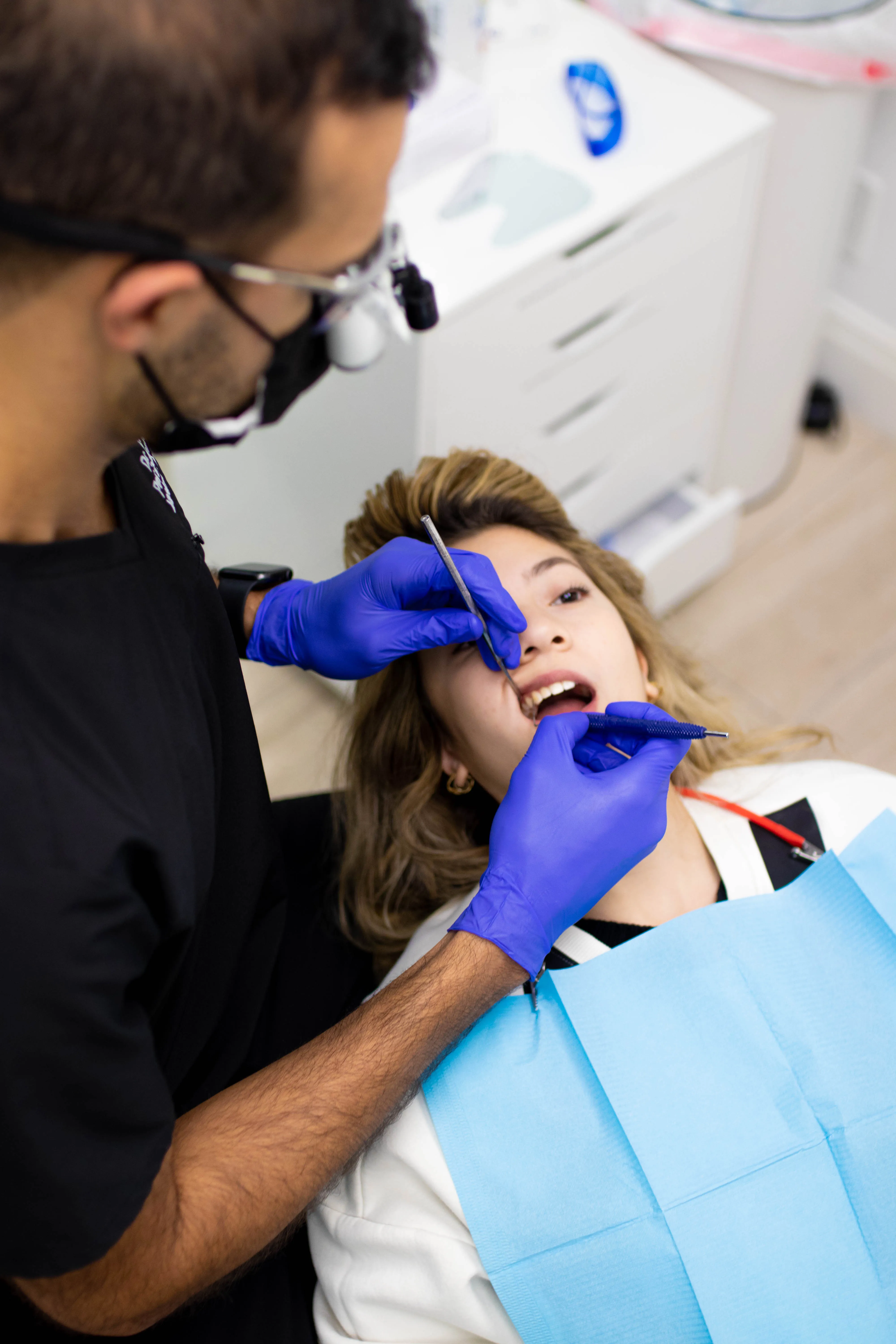 Dr. Rafik Dib wearing purple gloves examines a female patient's teeth using dental tools in a clinic.