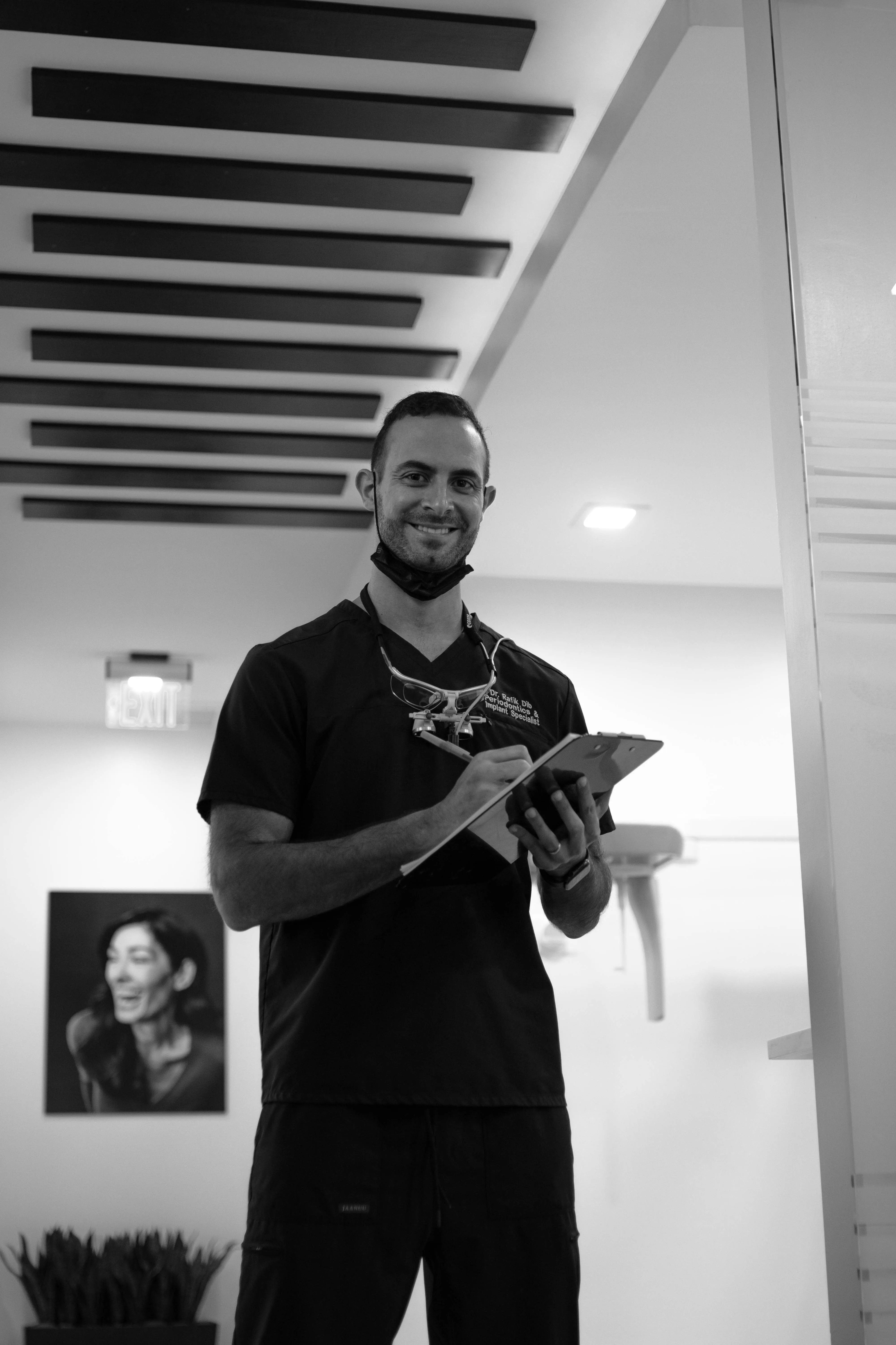 Dr. Rafik Dib wearing scrubs and a protective mask around his neck, holding a clipboard and pen in a dental office.