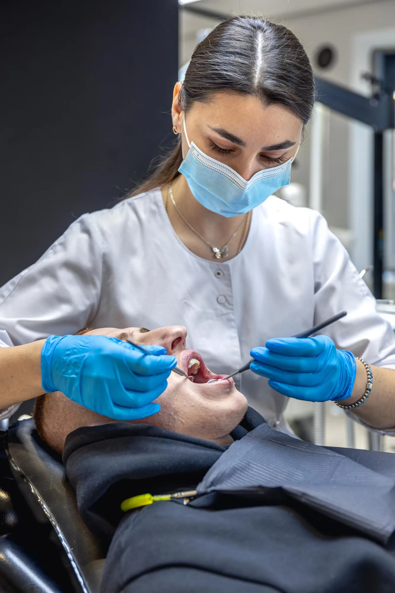 Dentist wearing blue gloves and a face mask examining a patient's teeth with dental tools.
