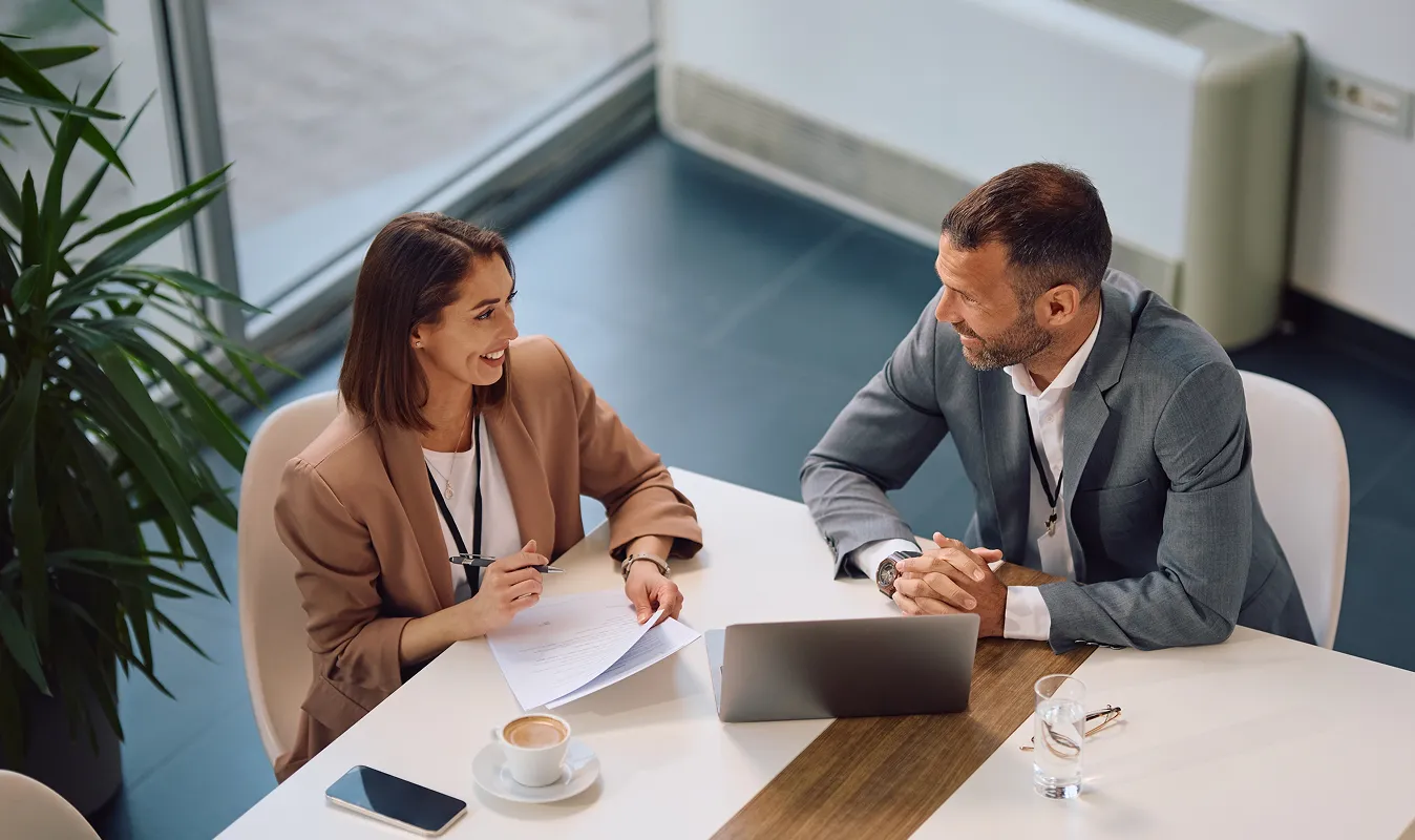 Two business professionals smiling during a meeting at a white table with laptop, coffee, and documents.