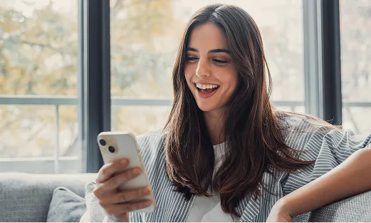 Smiling woman with long dark hair looking at her phone while sitting on a couch near large windows.