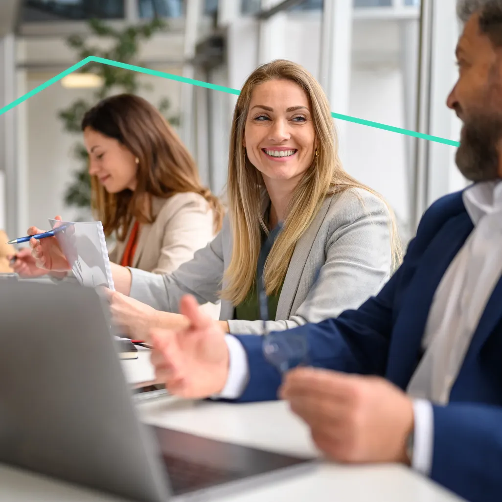 Three business professionals sitting at a table with laptops, engaged in a discussion, with a woman smiling at a male colleague.