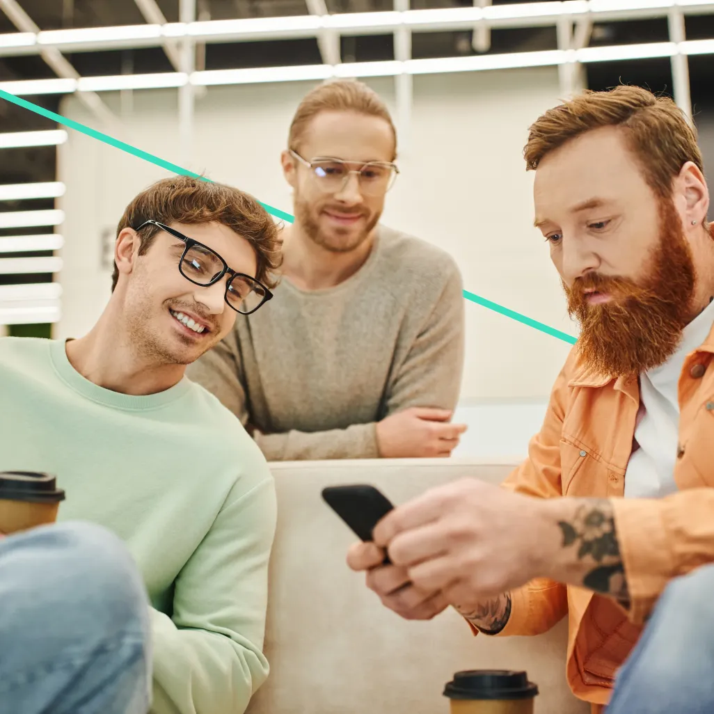 Three men with glasses and casual clothing sitting together, one showing something on a phone while others watch with interest.