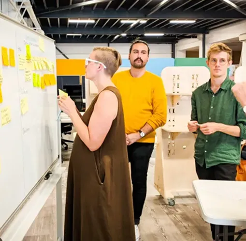 Three people collaborating in an office, one placing sticky notes on a whiteboard while two others observe.