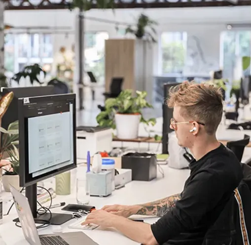 Person with tattoos wearing wireless earbuds working on a desktop computer in a modern office with plants and natural light.