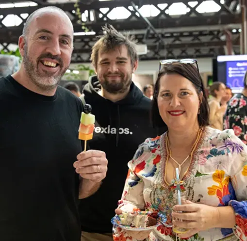 Three people smiling at a social event, one holding a fruit skewer, another holding a drink and plate of food.