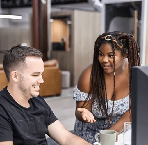 Two colleagues in a modern office, a woman explaining something to a smiling man seated at a desk with a computer.