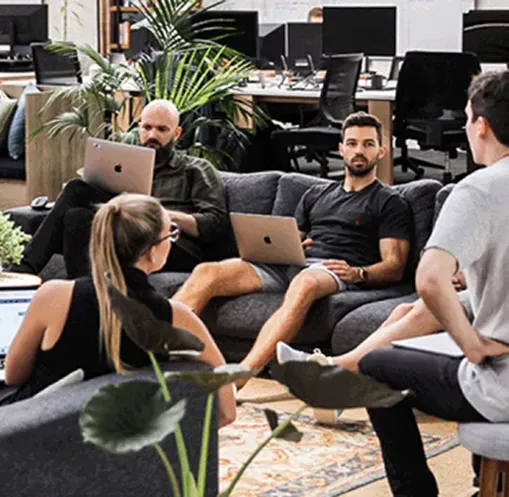 Four colleagues sitting in a modern office lounge area, engaged in discussion, with laptops on their laps.