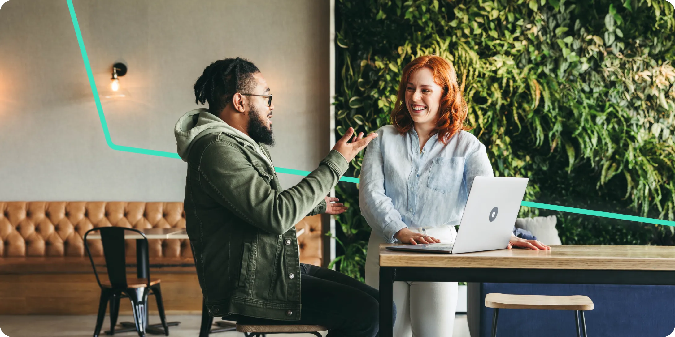 Two colleagues smiling and talking in a modern office with a laptop on the table and a green plant wall in the background.