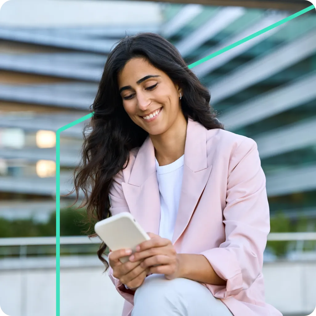 Smiling woman with long dark hair wearing a pink blazer, sitting outdoors and looking at a smartphone.