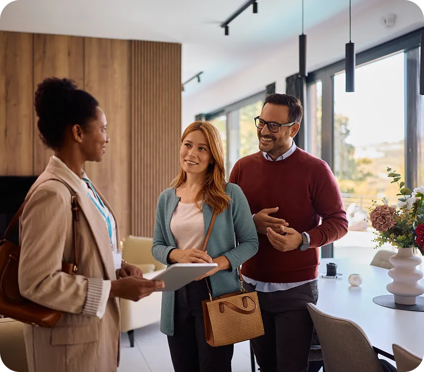 A real estate agent speaking with a smiling couple inside a modern, sunlit home.