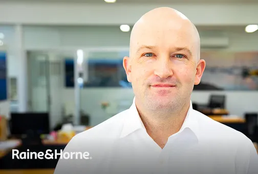 Portrait of a bald man in a white shirt smiling in an office setting with Raine & Horne logo.