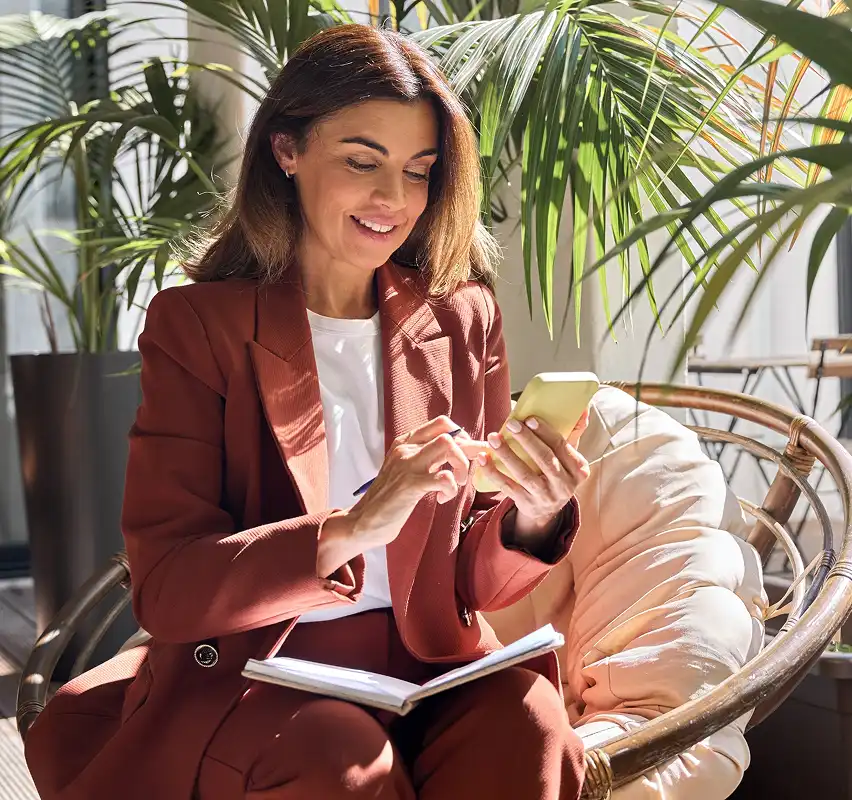 Smiling woman in brown suit sitting on a cushioned chair, using a smartphone with a notebook on her lap, surrounded by indoor plants.