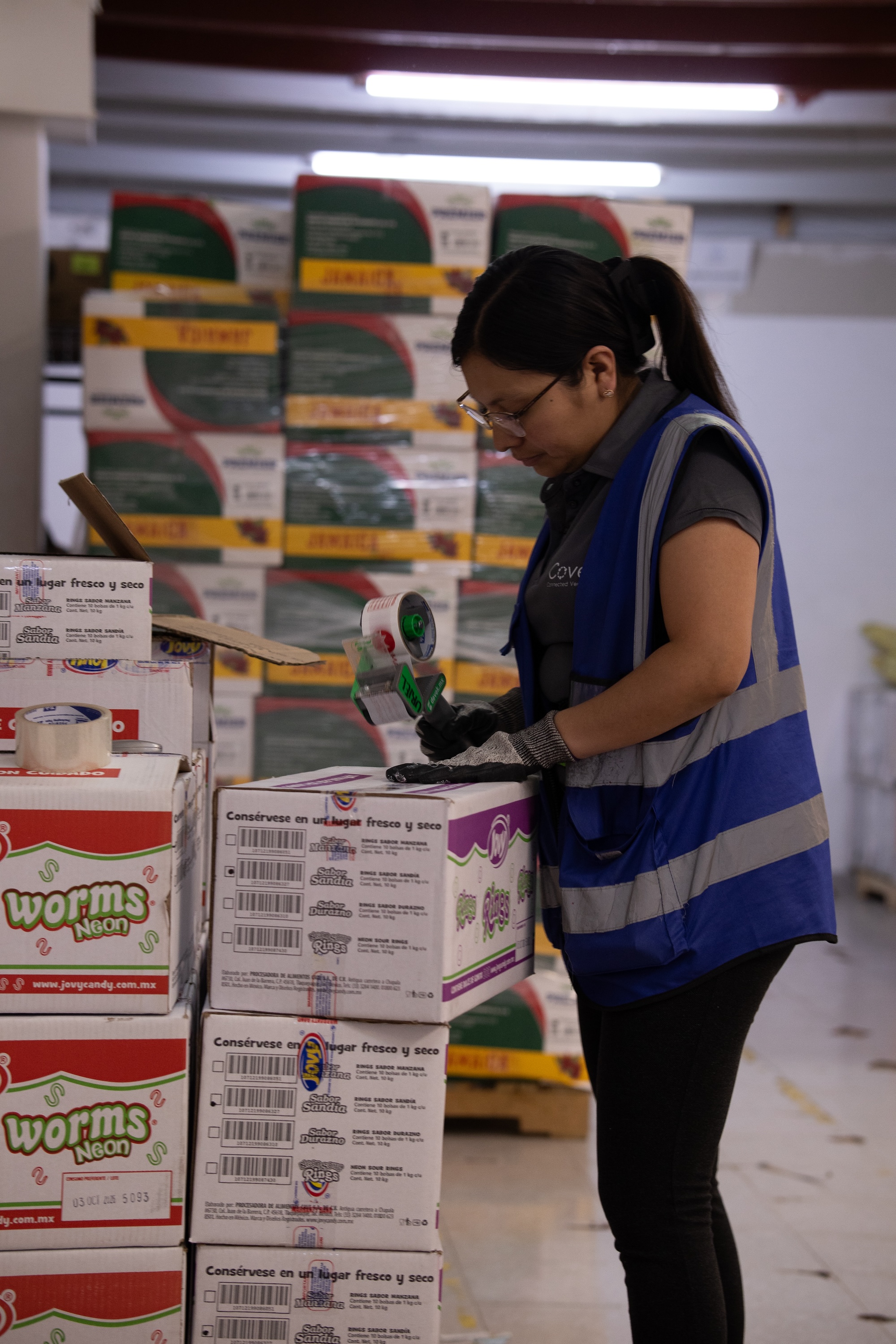 Mujer con chaleco azul y guantes empaqueta cajas de dulces en un almacén.