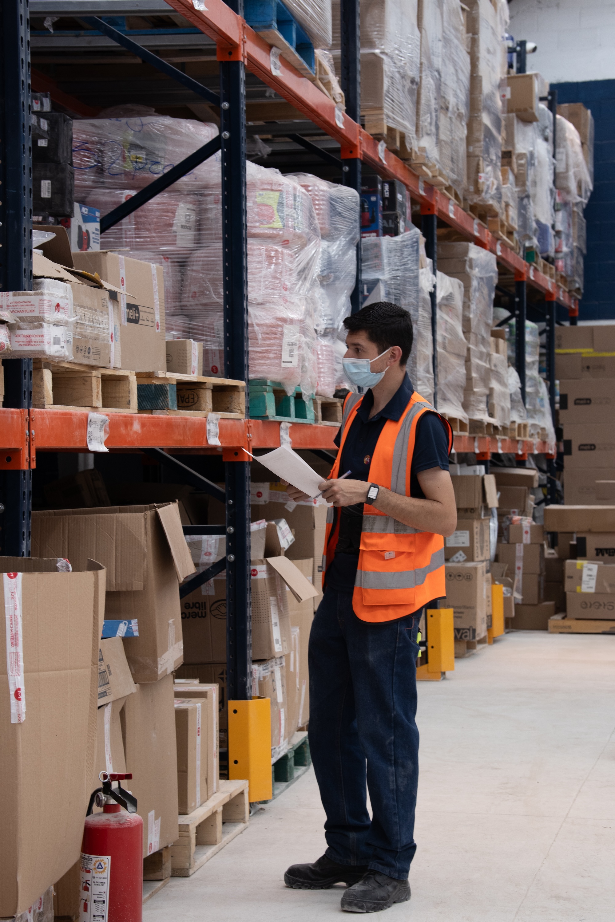 Trabajador con chaleco reflectante y cubrebocas revisa inventario en bodega con estanterías llenas de cajas y pallets.