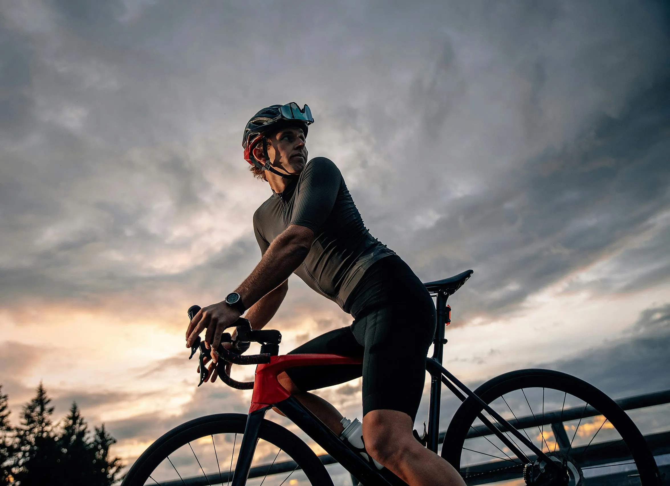 Cyclist wearing a helmet and black cycling outfit on a red and black road bike during a cloudy sunset.