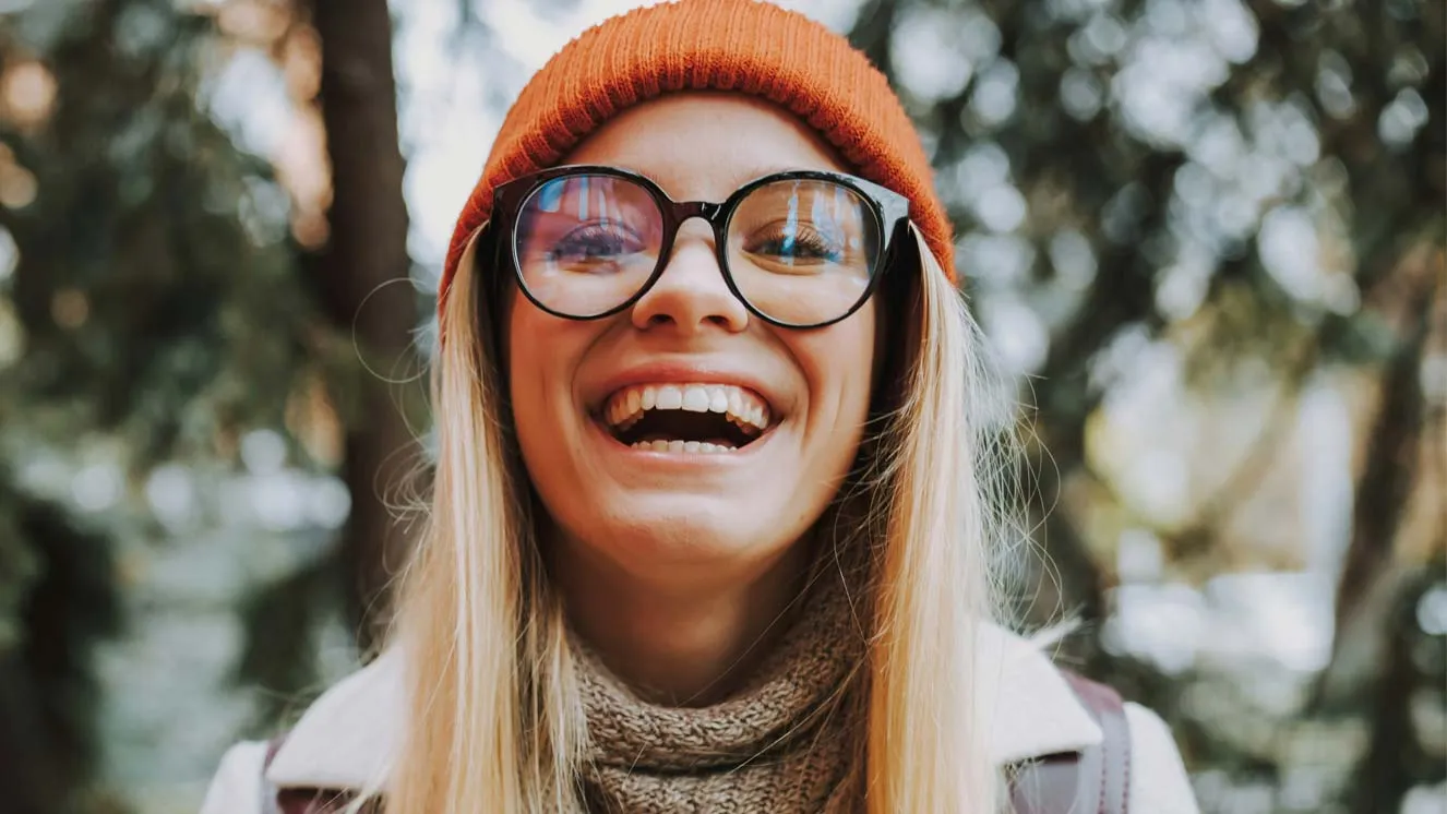 Smiling woman wearing large black glasses, an orange beanie, and a brown scarf outdoors.