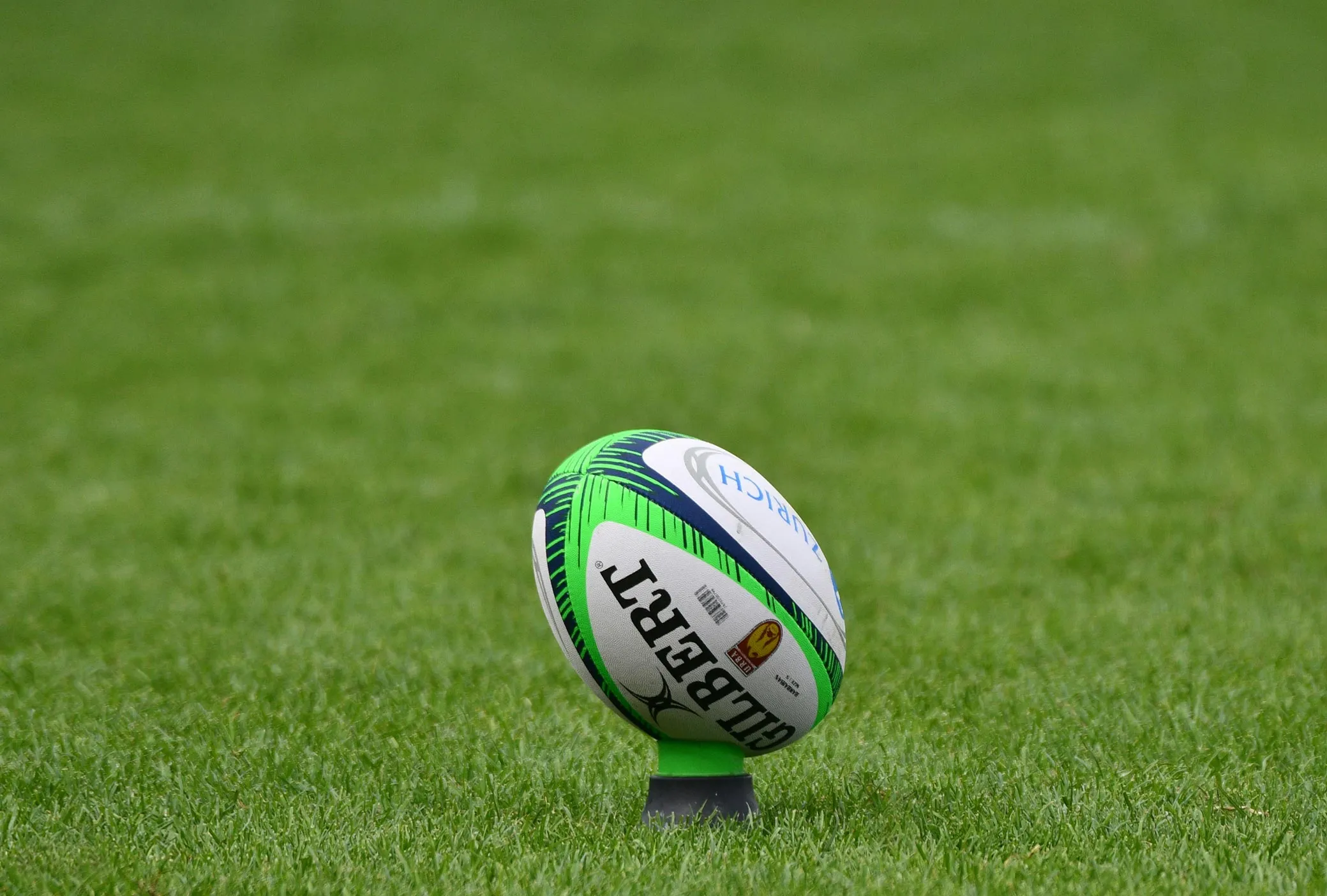 A Gilbert rugby ball standing upright on a kicking tee on a grassy field.