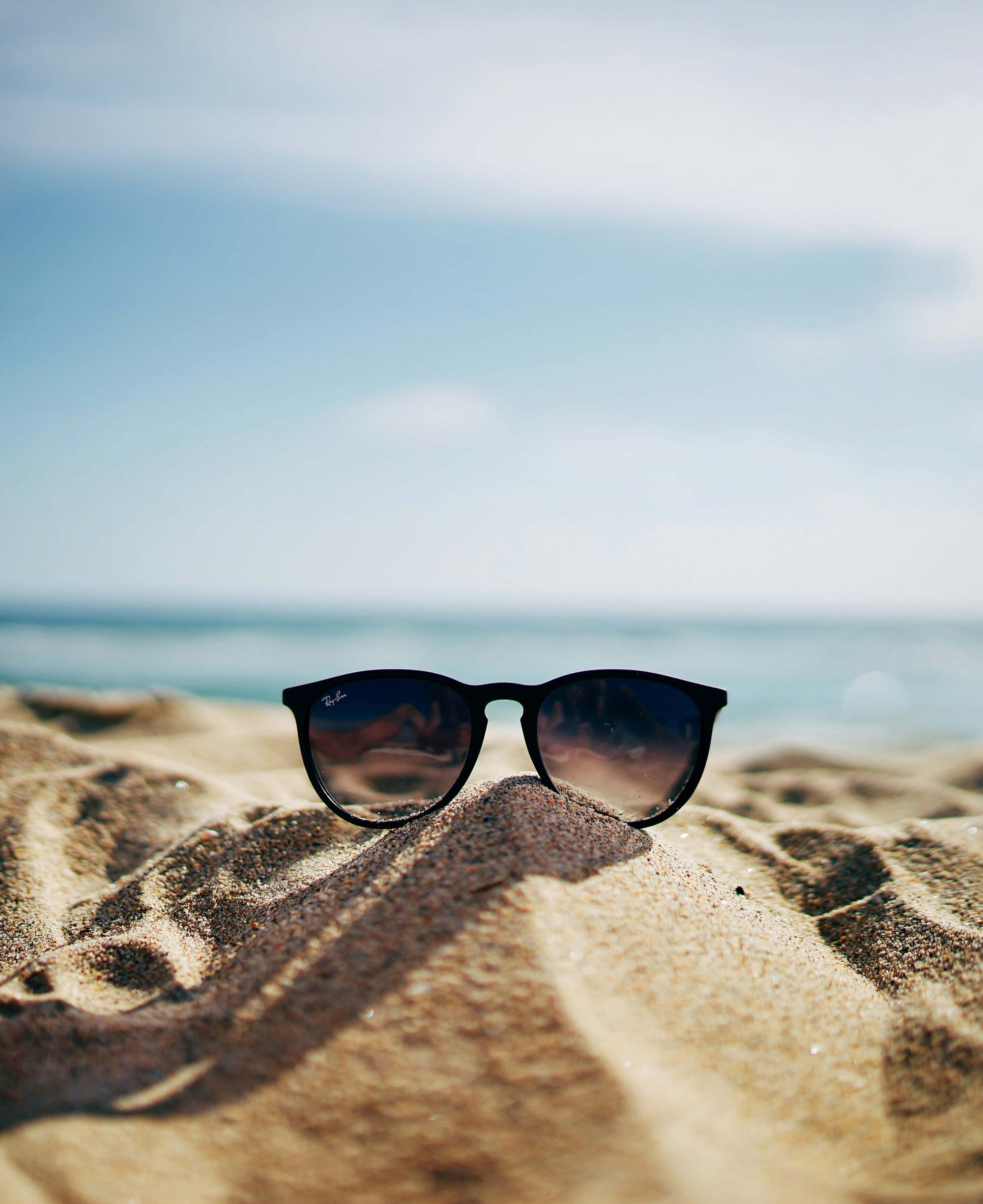 Black Ray-Ban sunglasses resting on a small sand mound at the beach with the ocean and sky in the background.