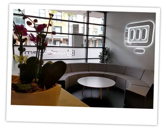Modern waiting area with curved grey sofa, round white coffee table, indoor plant, and illuminated logo on the wall.