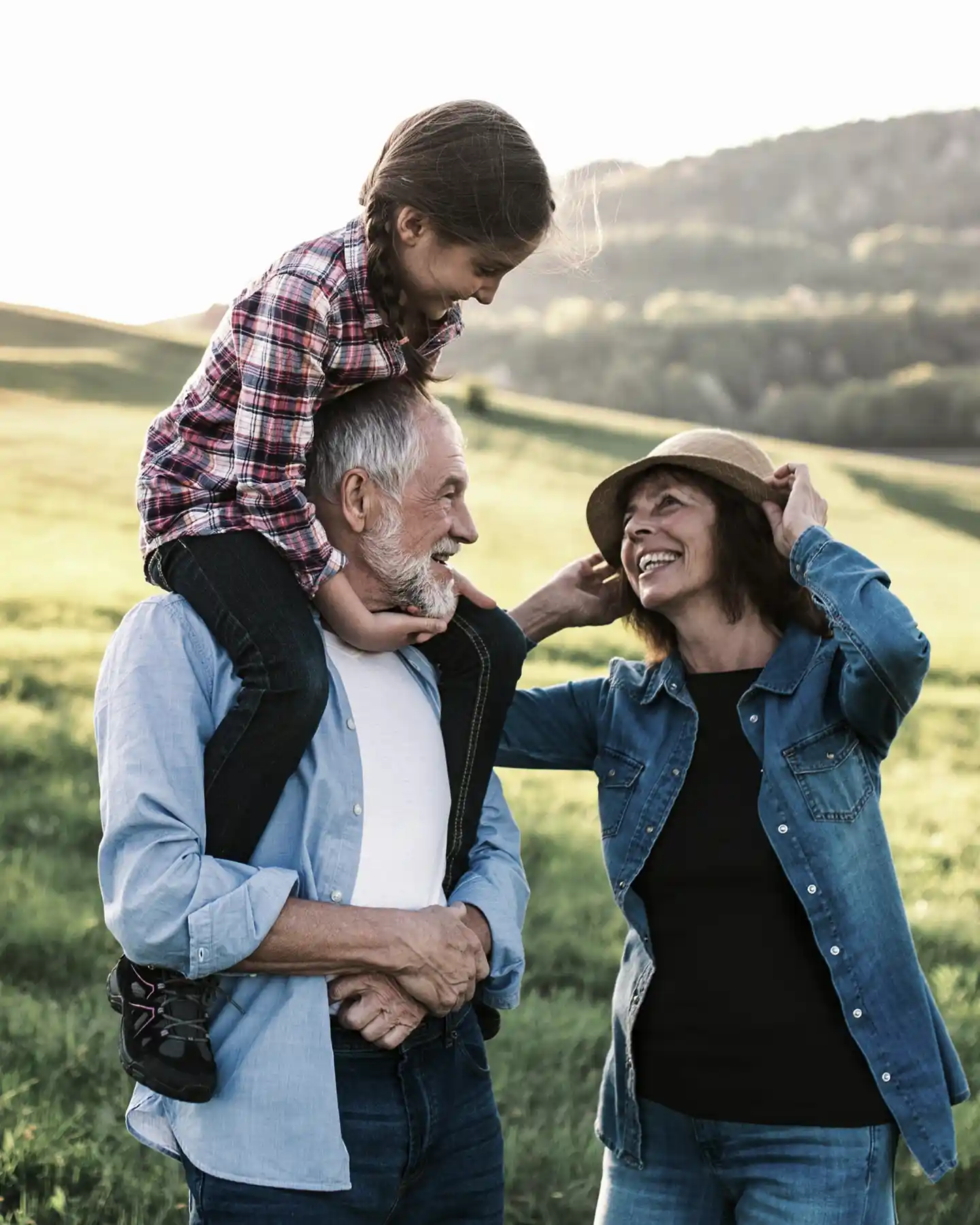Smiling elderly man carrying a young girl on his shoulders while an elderly woman adjusts her hat outdoors in a green field.