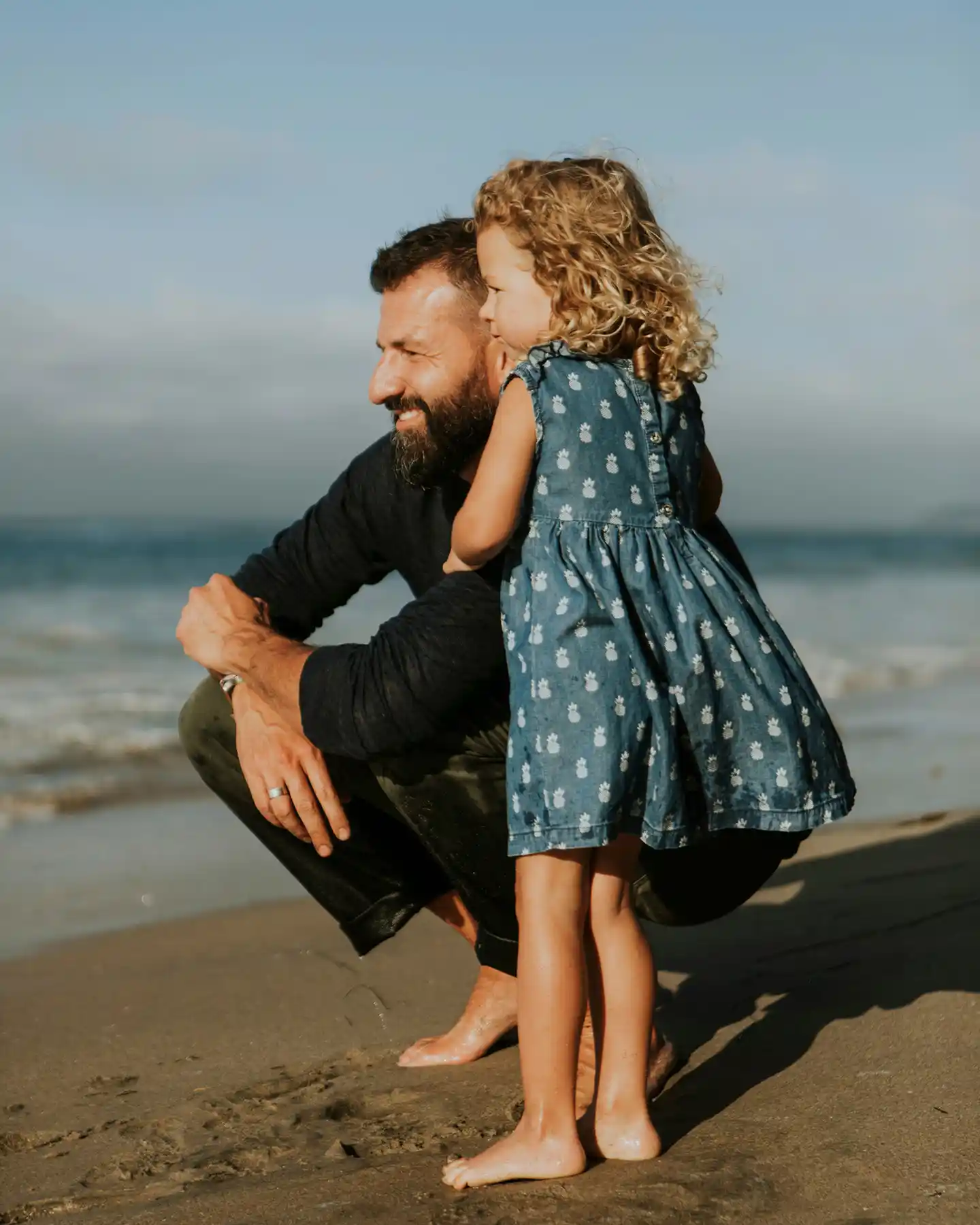 Man crouching on a sandy beach beside a young girl wearing a blue dress with a pineapple pattern, both barefoot.