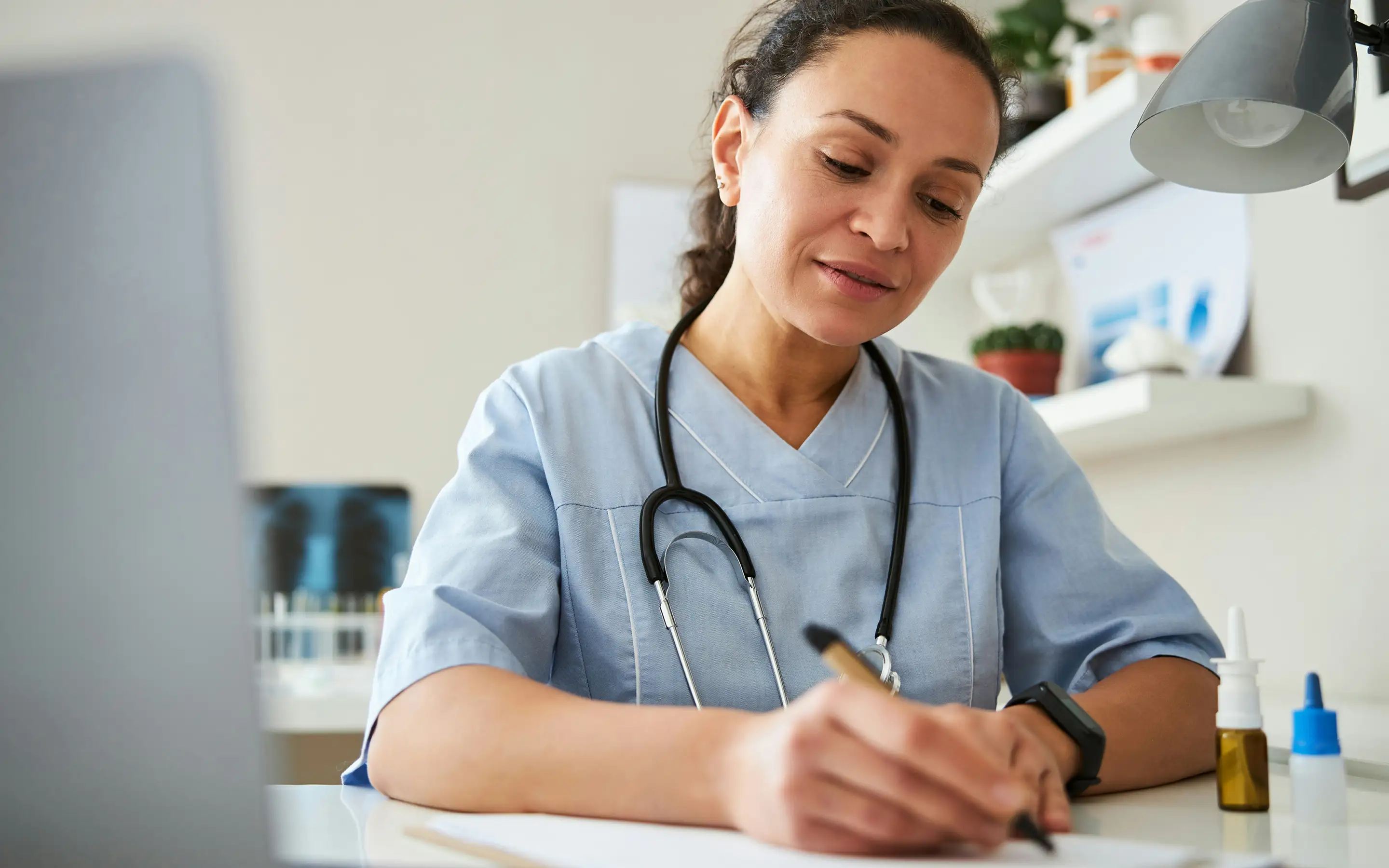Female healthcare professional in blue scrubs writing on paper with a stethoscope around her neck.