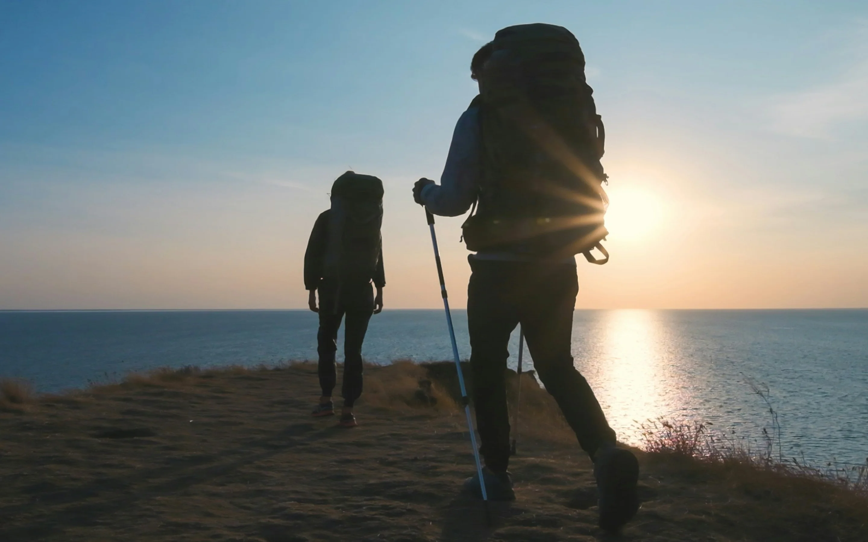 Two hikers with large backpacks walking on a coastal trail at sunset with the sun reflecting on the sea.