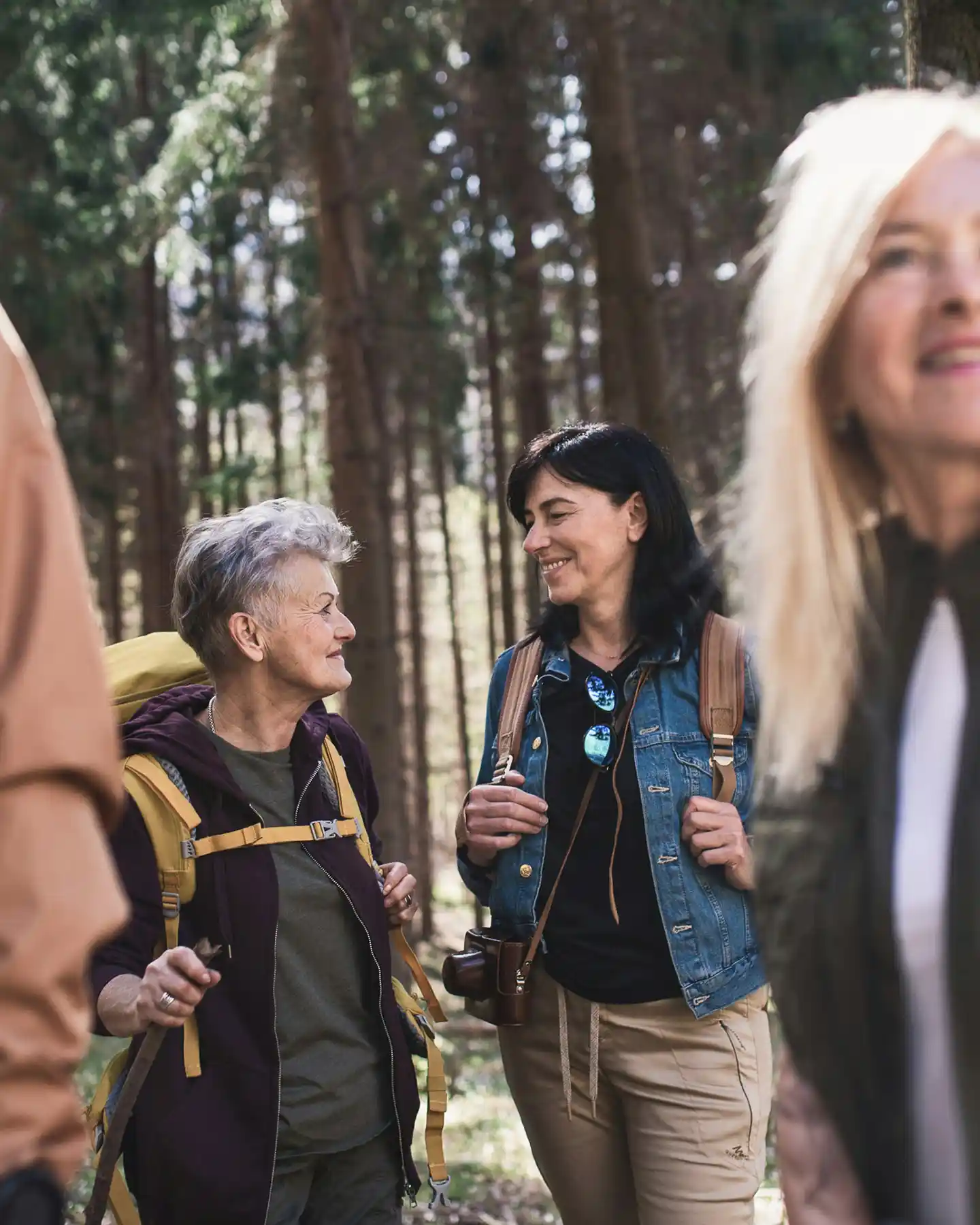Two women smiling and talking while hiking in a forest with backpacks.