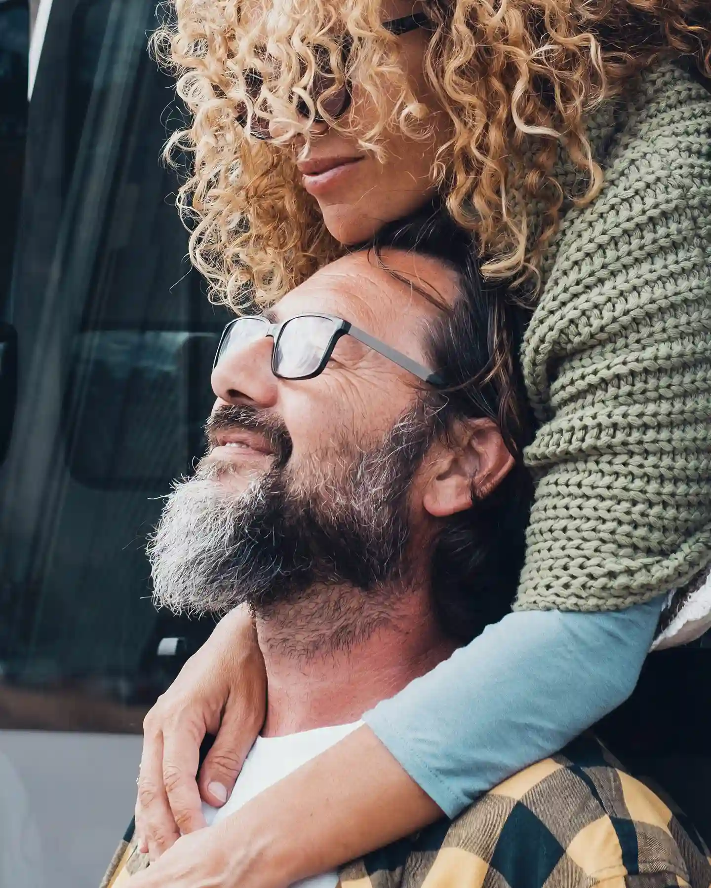 Curly-haired woman embraces smiling bearded man wearing glasses and plaid shirt.