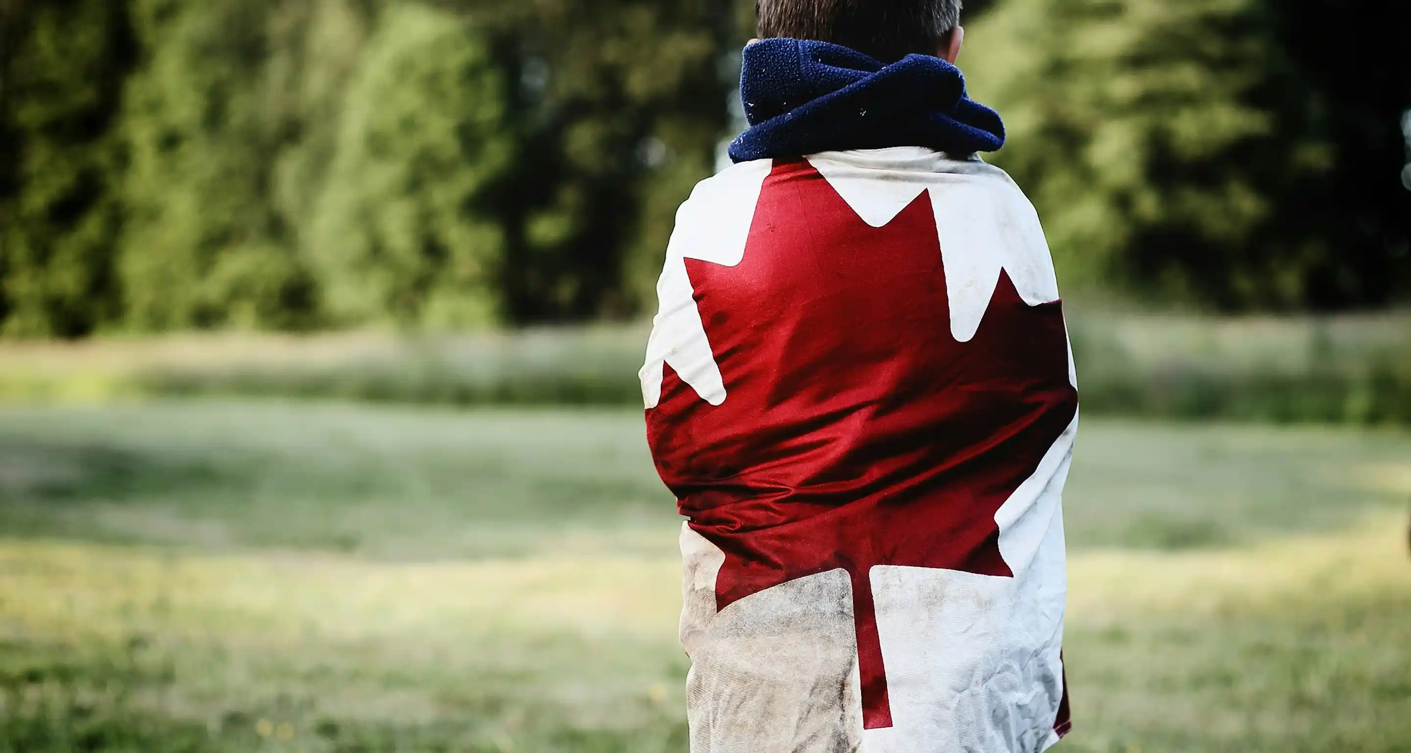 Person wrapped in a Canadian flag blanket standing outdoors with a blurred green background.