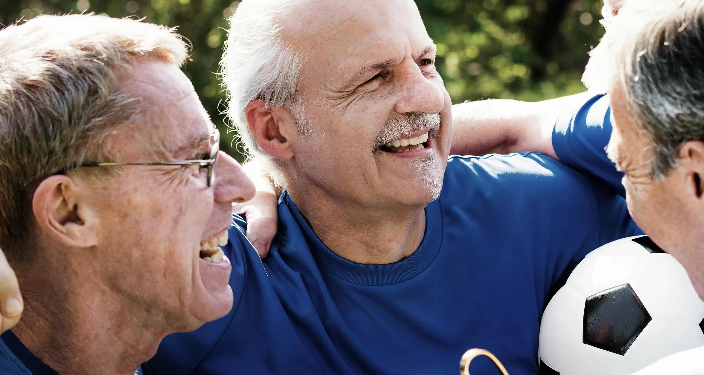 Three older men in blue shirts joyfully embracing each other outdoors with a soccer ball.