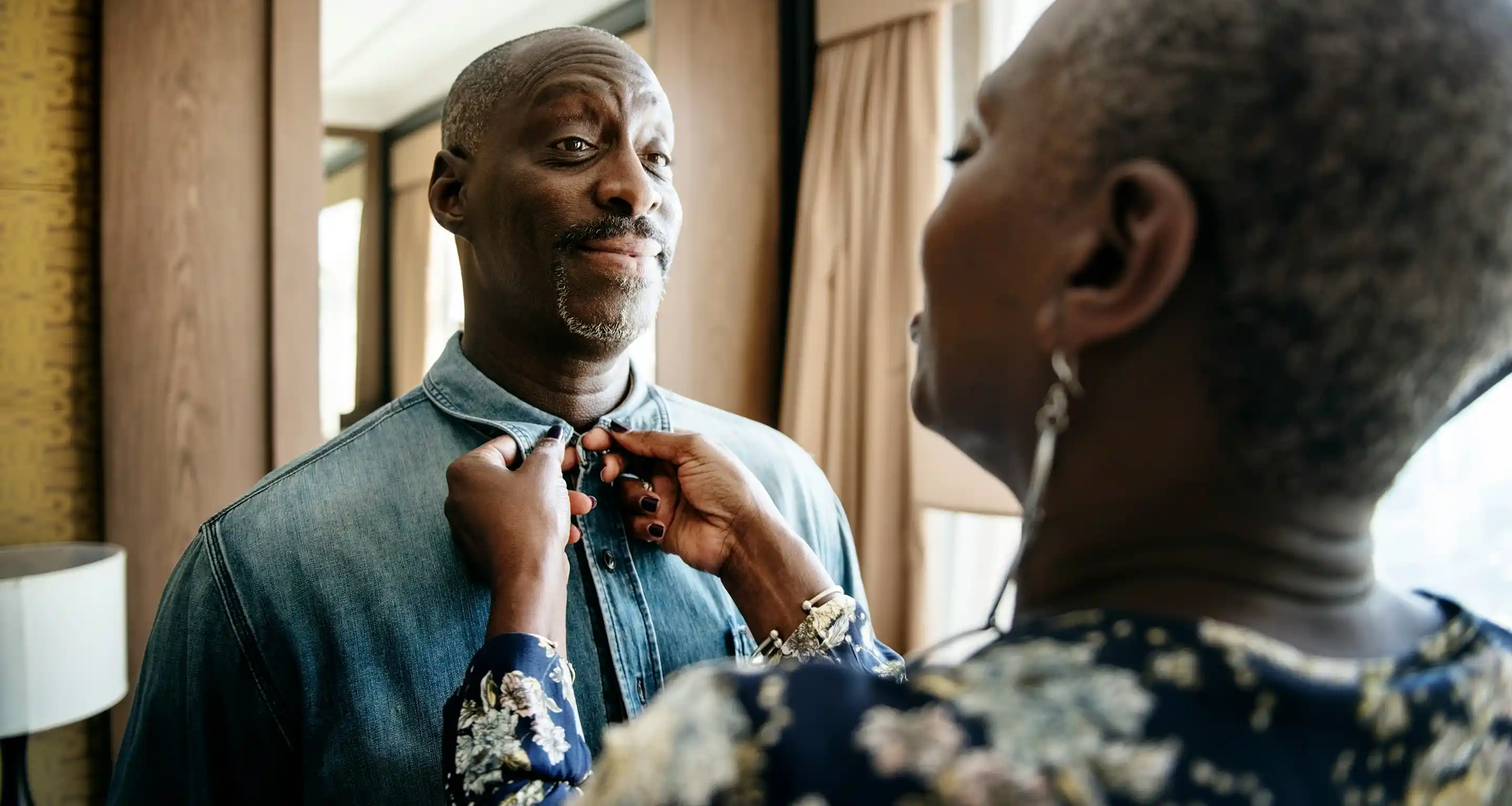 Person fastening the top button of a denim shirt worn by another person indoors near a window.