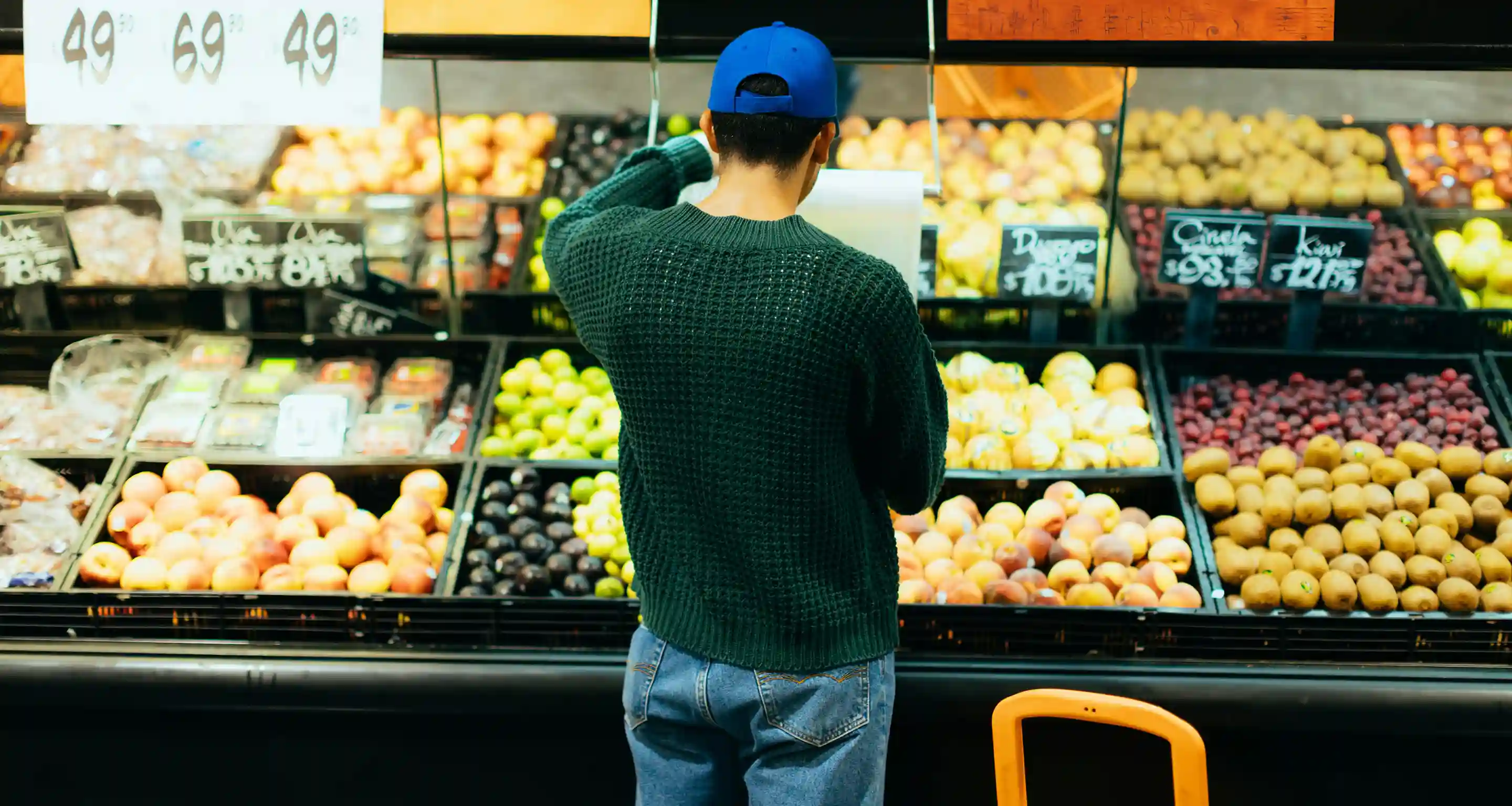 Person wearing a blue cap and green sweater selecting fruit in a grocery store produce section.