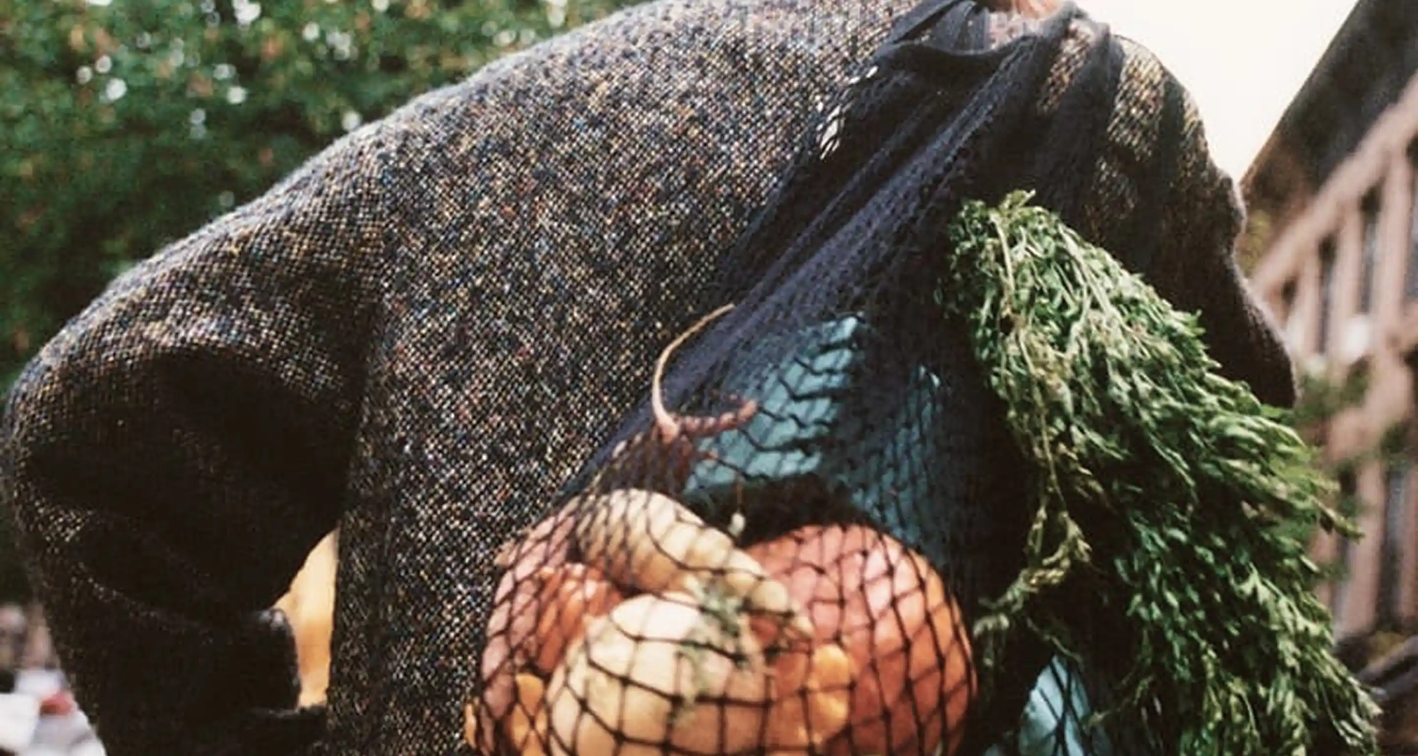 Person carrying a black mesh bag filled with root vegetables and fresh green herbs over their shoulder.