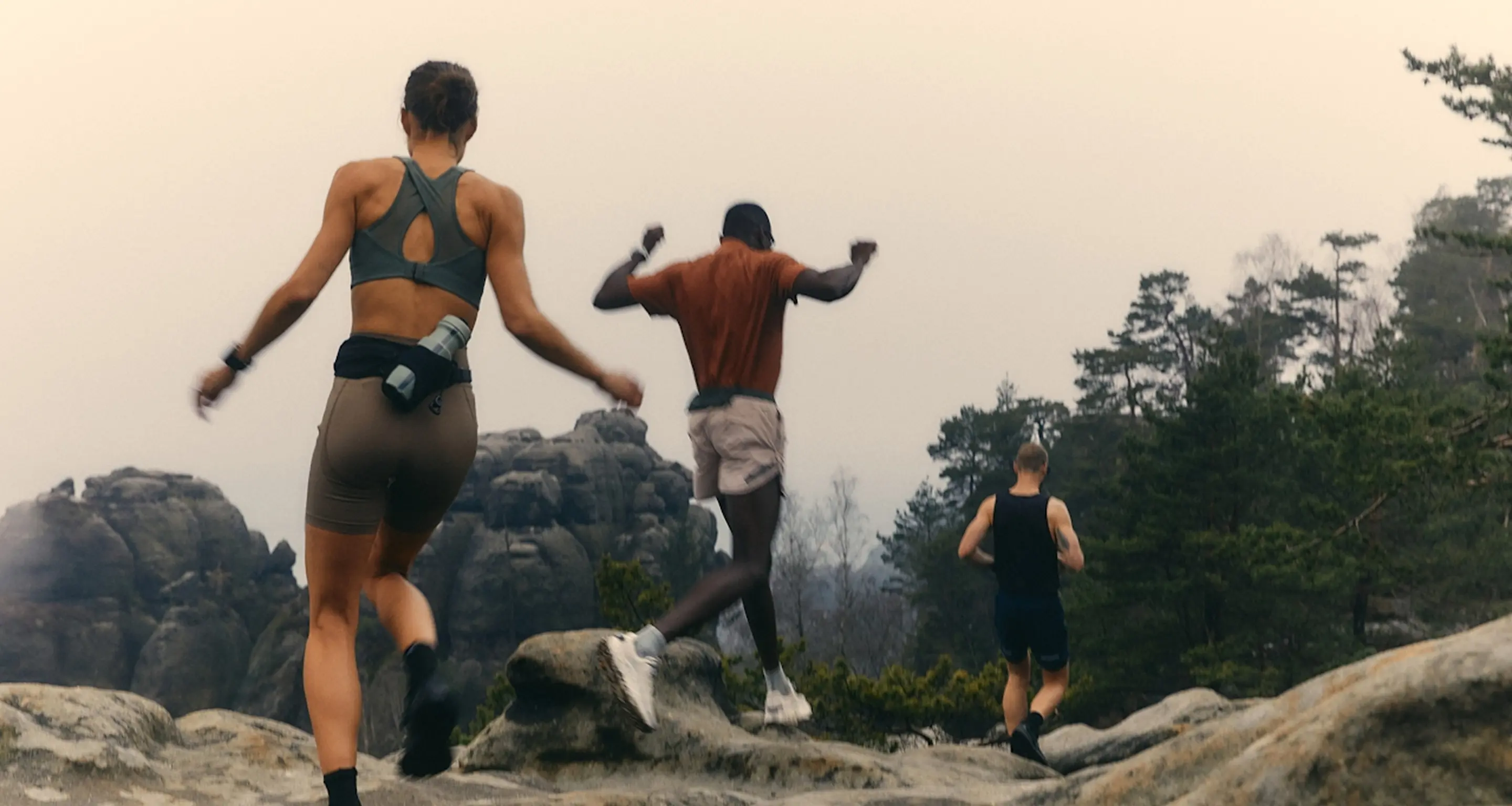 Three people running and jumping over rocky terrain in an outdoor forest setting.