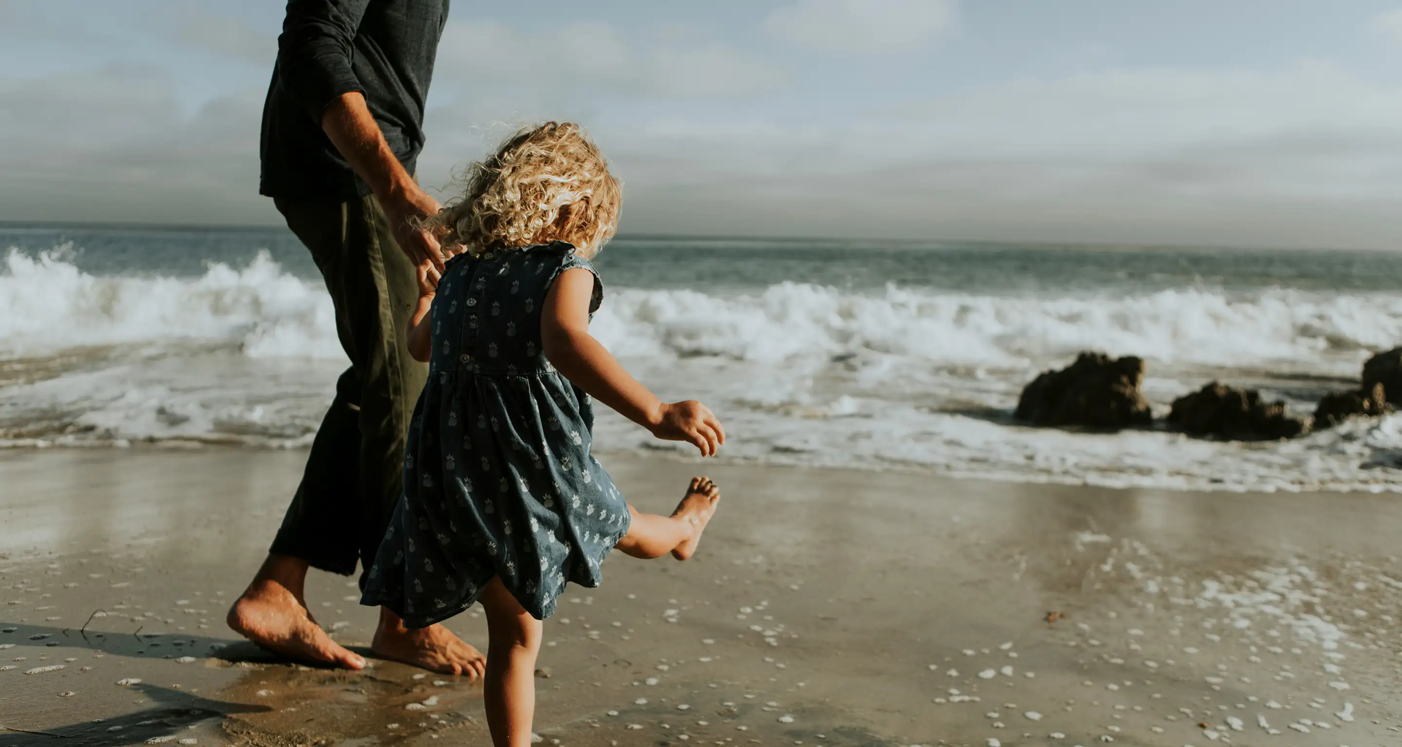 Young girl in a blue dress holding hands with an adult walking barefoot on the beach near ocean waves.