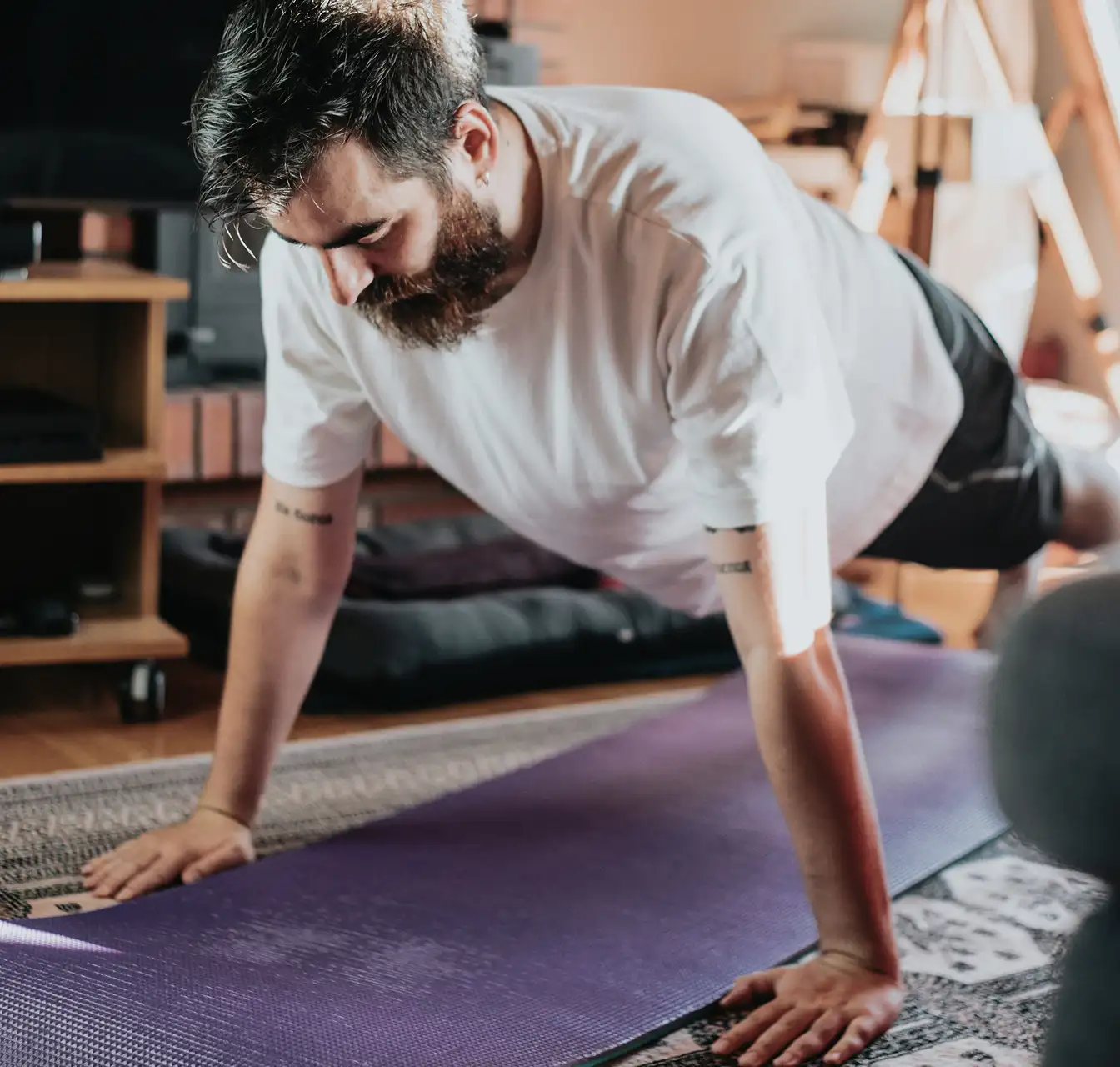Bearded man in a white t-shirt doing a push-up on a purple yoga mat indoors.