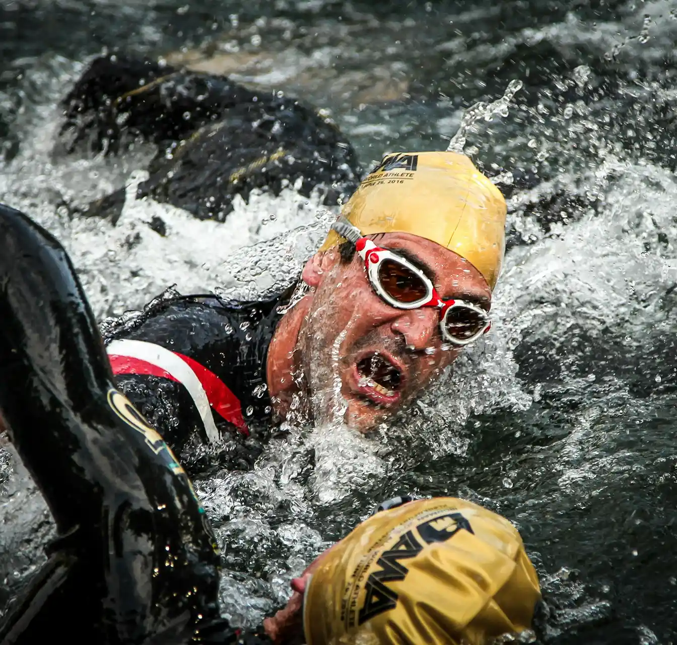Swimmer wearing a yellow cap and white goggles emerging from water with splashes around him.