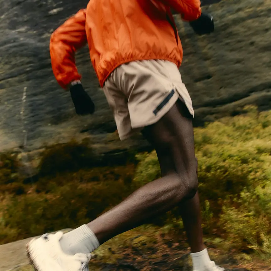Person in orange jacket and beige shorts running outdoors on a mossy and rocky terrain.