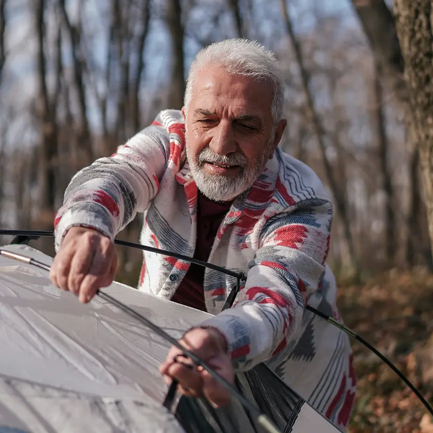 Smiling elderly man assembling poles for a white camping tent in a forest.