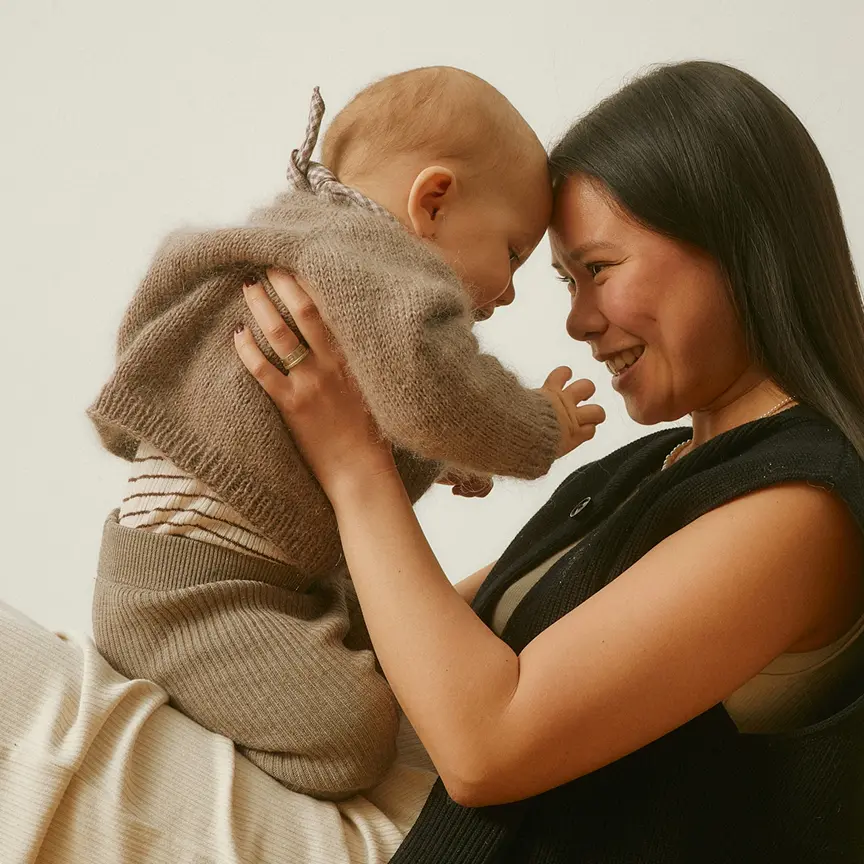 Woman smiling and holding a baby who is reaching out to her, both dressed in cozy knitwear.