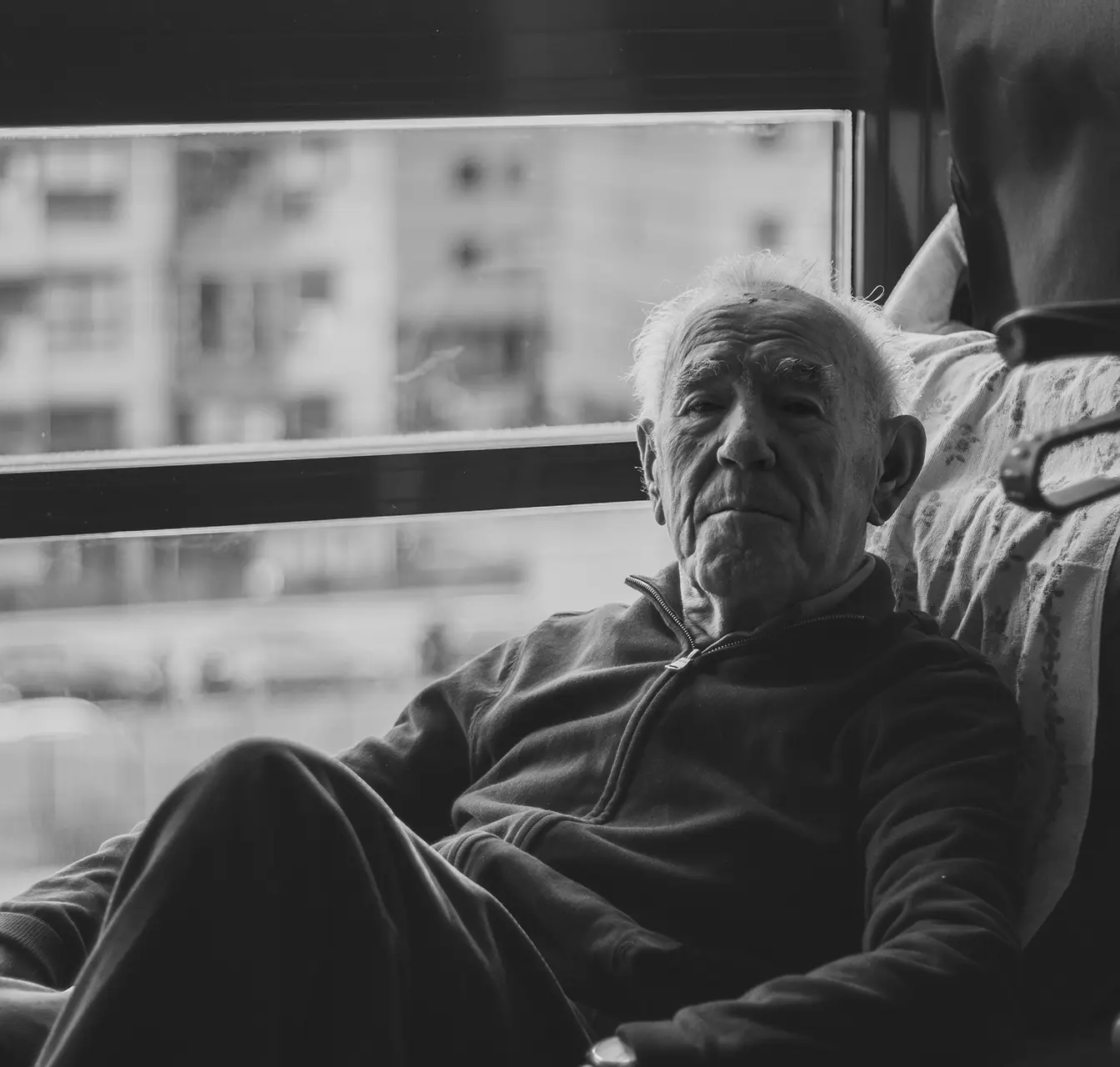 Black and white portrait of an elderly man sitting indoors near a window with a contemplative expression.