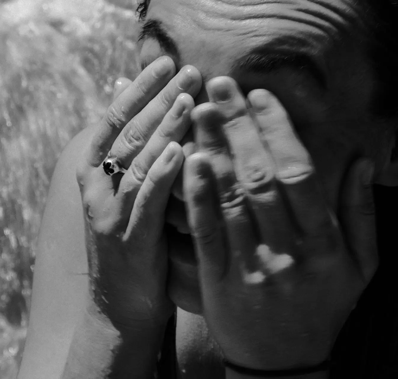 Close-up black and white photo of a person covering their face with both hands, wearing a ring with a dark stone on the left hand.