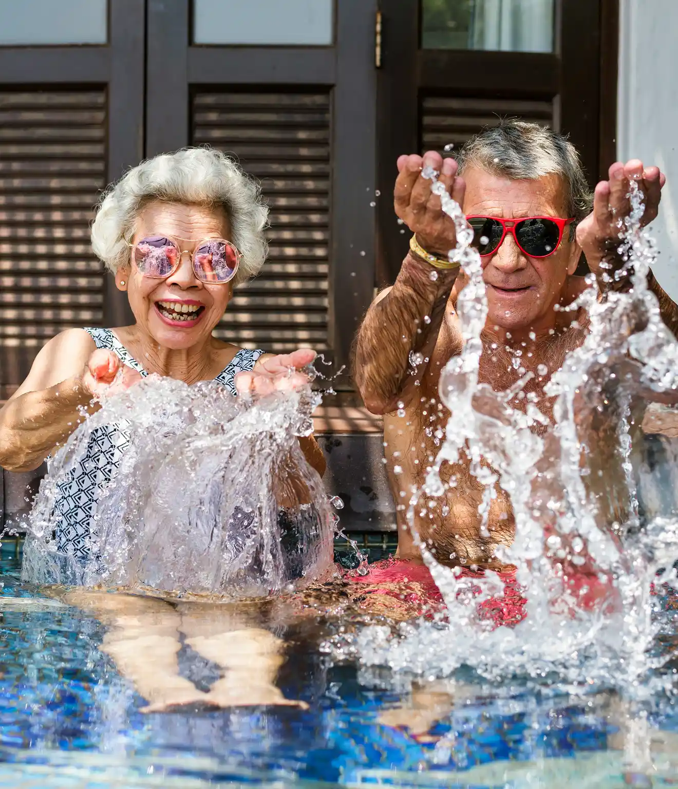 Senior couple wearing sunglasses playfully splashing water while sitting in a pool.
